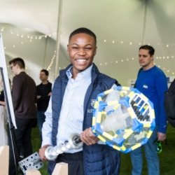 A person smiling broadly holds a large, colorful geometric object with clear panels in an event tent. Other attendees are visible in the background, engaged in activities and discussions. The tent is decorated with string lights.